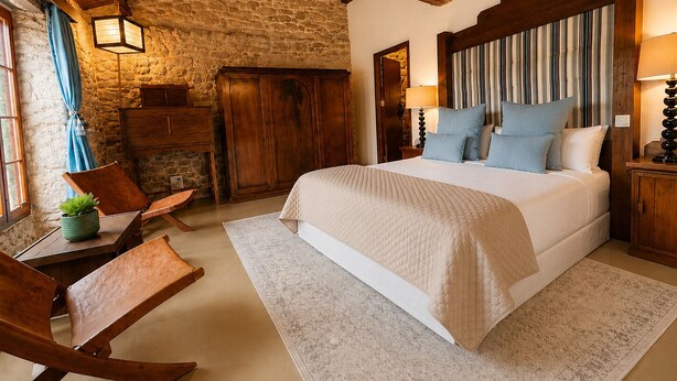 A bedroom with a stone wall, wooden beams, a bed with pillows, a nightstand, a lamp, and a chair.