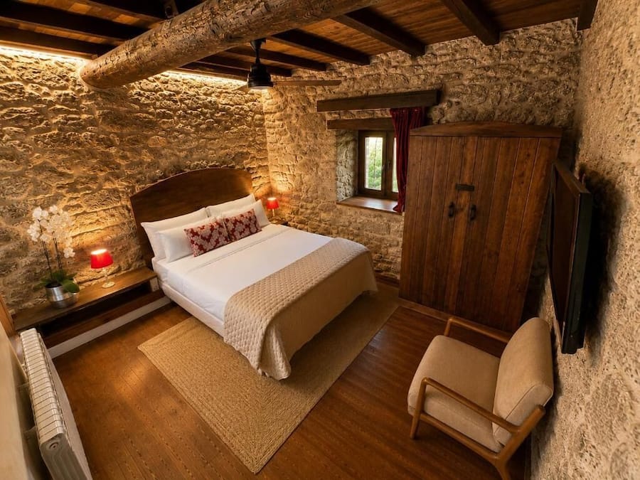 Un dormitorio con pared de piedra, una cama de madera, una mesita de noche con lámpara, un armario de madera y una ventana con cortinas.