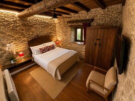 A bedroom with a stone wall, a wooden bed, a nightstand with a lamp, a wooden wardrobe, and a window with curtains.