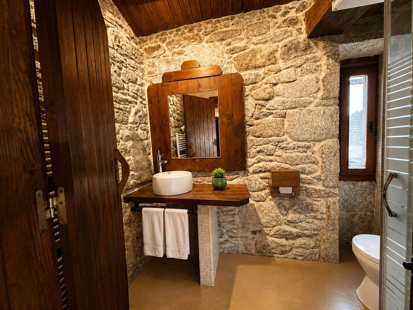 A bathroom with a stone wall, wooden vanity, a sink, a mirror, a potted plant, and a toilet.