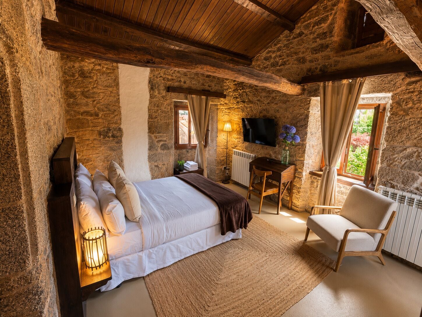 Un dormitorio rústico con pared de piedra, ropa de cama blanca, un escritorio de madera, una silla y un televisor.