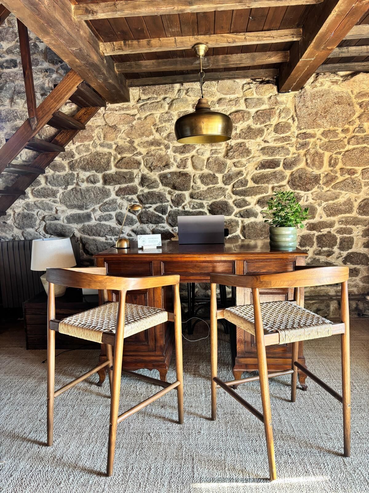 Una mesa de madera rústica con dos sillas, un candelabro de latón colgante y una pared de piedra al fondo.