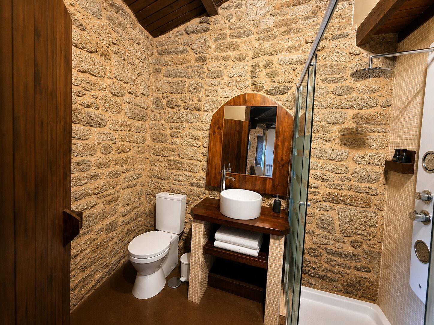 A bathroom with a stone wall, a wooden vanity, a white sink, a toilet, and a glass shower enclosure.