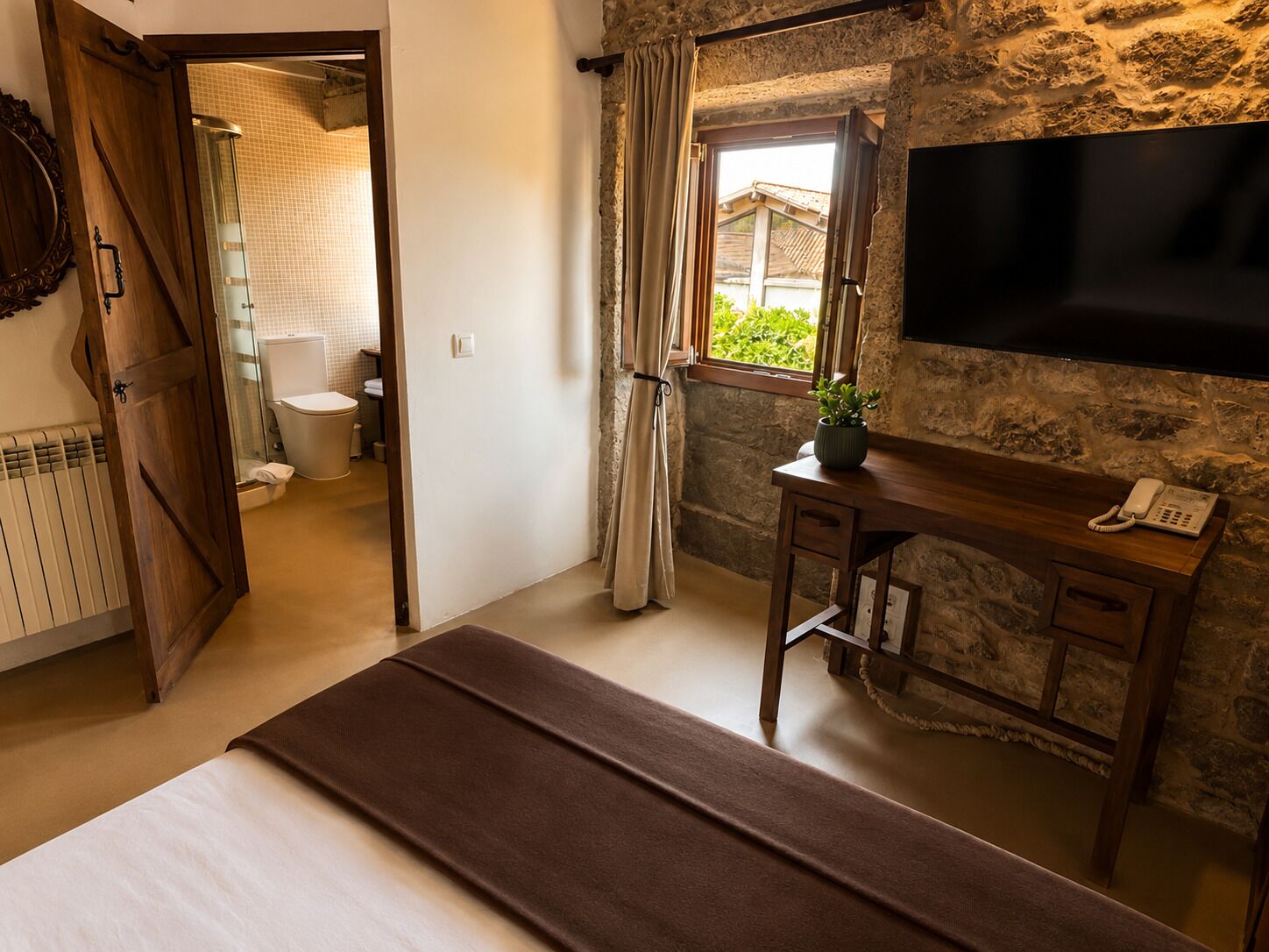 Una habitación de hotel con cama, un escritorio con teléfono, una pared de piedra, una ventana con cortinas y un baño visible a través de una puerta abierta.