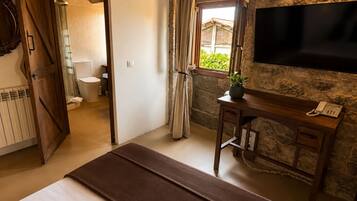 Una habitación de hotel con cama, un escritorio con teléfono, una pared de piedra, una ventana con cortinas y un baño visible a través de una puerta abierta.