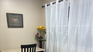 Ein Zimmer mit Holzboden, einem Tisch mit Stuhl, einem gerahmten Bild an der Wand, einer Vase mit Blumen und einem weißen Vorhang.