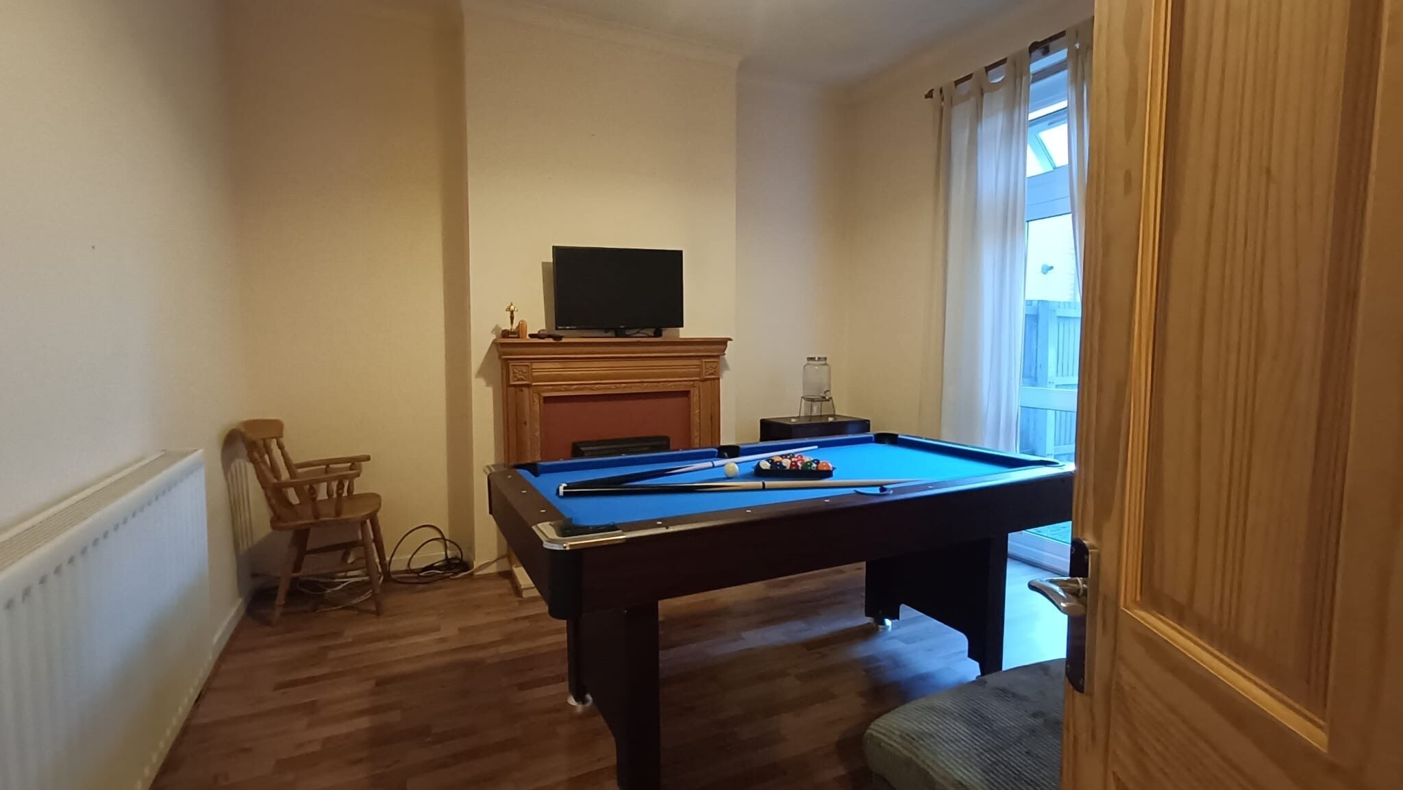 A room with a pool table, a television on a mantelpiece, a chair, and a window with curtains.