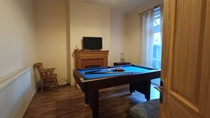 A room with a pool table, a television on a mantelpiece, a chair, and a window with curtains.