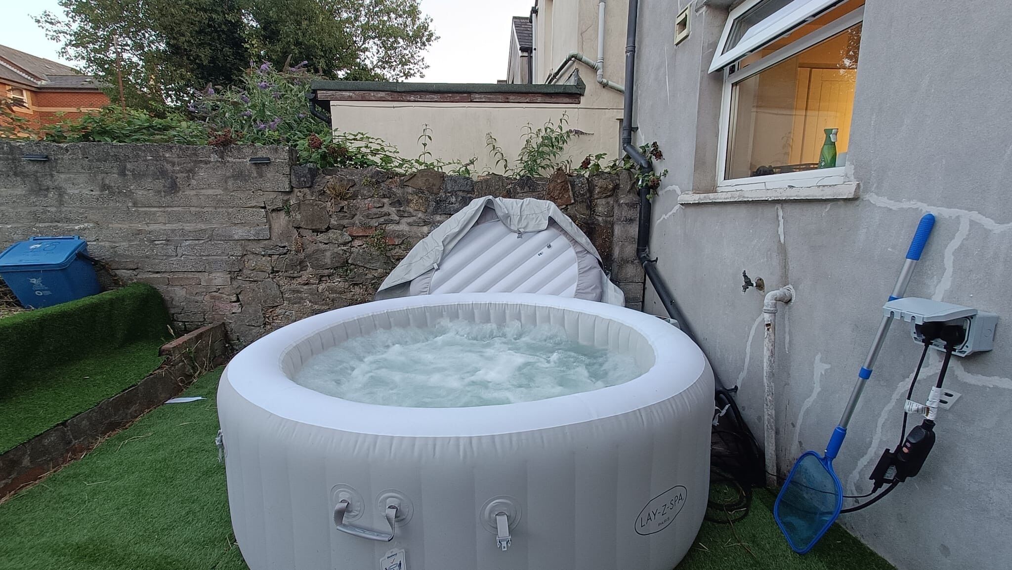 An inflatable hot tub in a backyard with a stone wall, a window, and a hose.