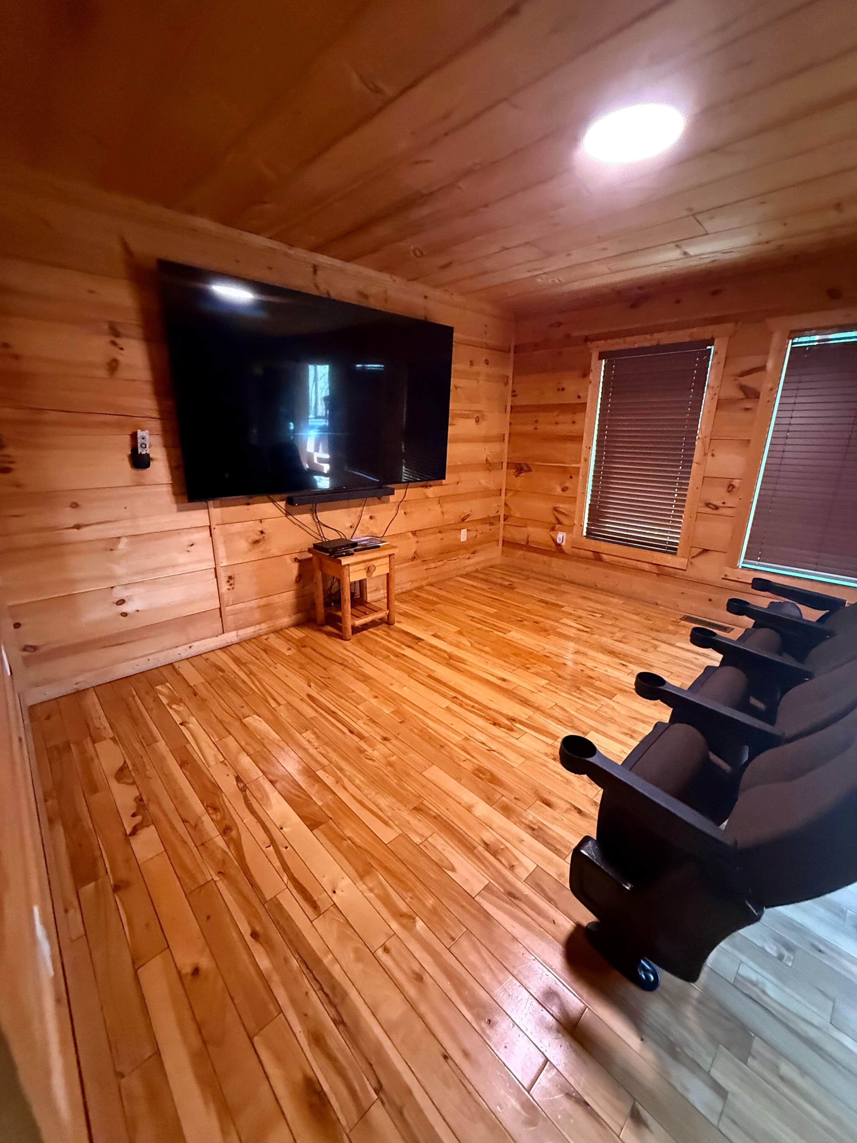Ein Zimmer mit Holzwandverkleidung, einem an der Wand befestigten Flachbildfernseher, einem kleinen Holztisch und einem Liegestuhl.