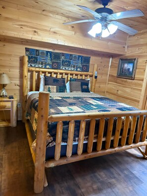 Ein Zimmer mit Holzboden, einem Bett, einem Nachttisch, einer Lampe und einem Deckenventilator.