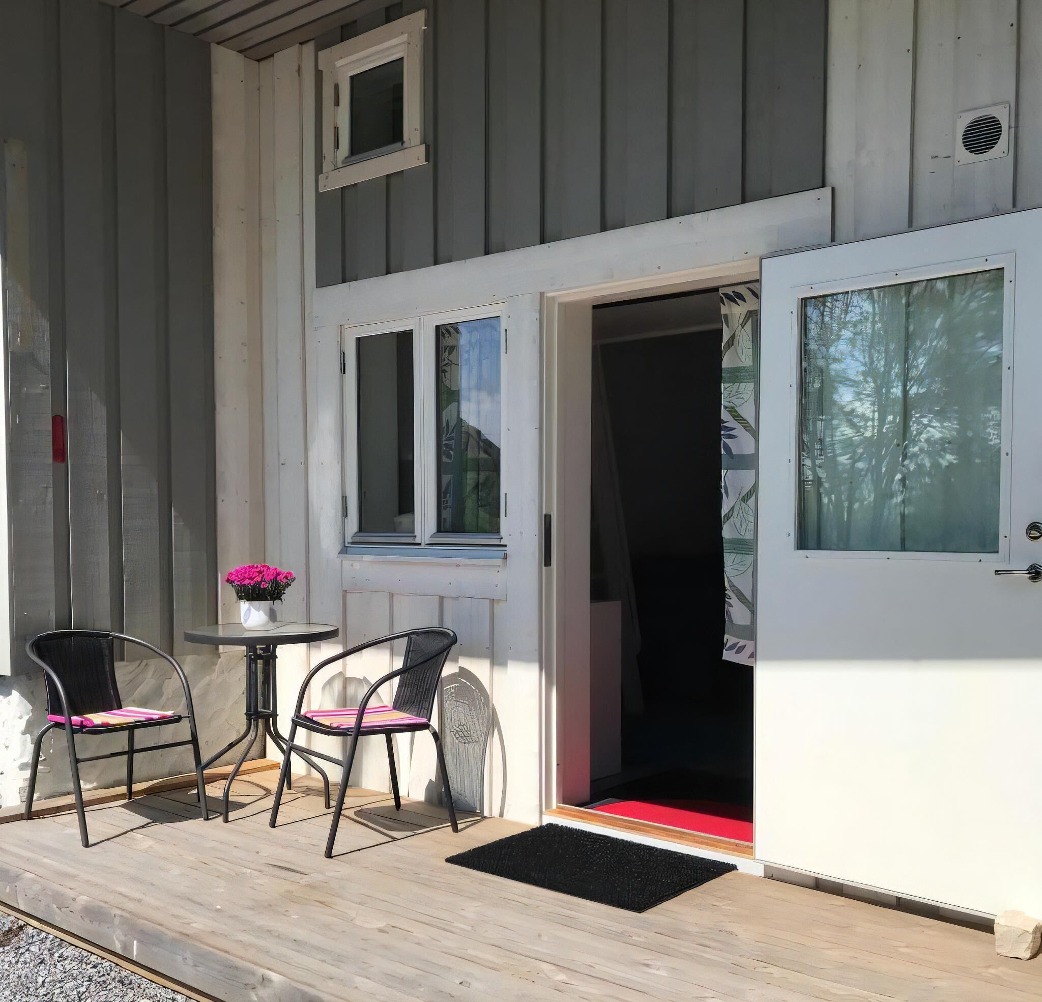 En träveranda med två stolar och ett litet bord, en dörr med en röd dörrmatta och ett fönster med en gardin.