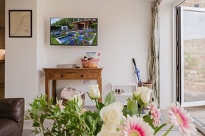A living room with a television displaying a garden scene, a wooden table with a basket, and a view of a brick wall through a window.
