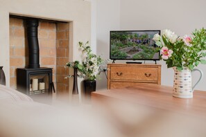 Ett vardagsrum med en öppen spis, en tv-skärm som visar en bild av en trädgård och en vas med blommor på ett träbord.