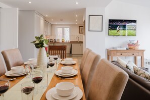 A dining area with a long table set for a meal, a television displaying a sports event, and a kitchen in the background. - Kempsey House -  Frampton on Severn, Gloucester, Tewkesbury (Frampton on Severn)