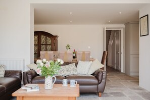 A living room with a brown leather sofa, a wooden coffee table with books and a vase of flowers, and a dining area with a wooden cabinet and a table set for two.