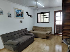 A living room with a dark brown sofa, a beige sofa, a ceiling fan, a window with blinds, and a door with a wooden frame.