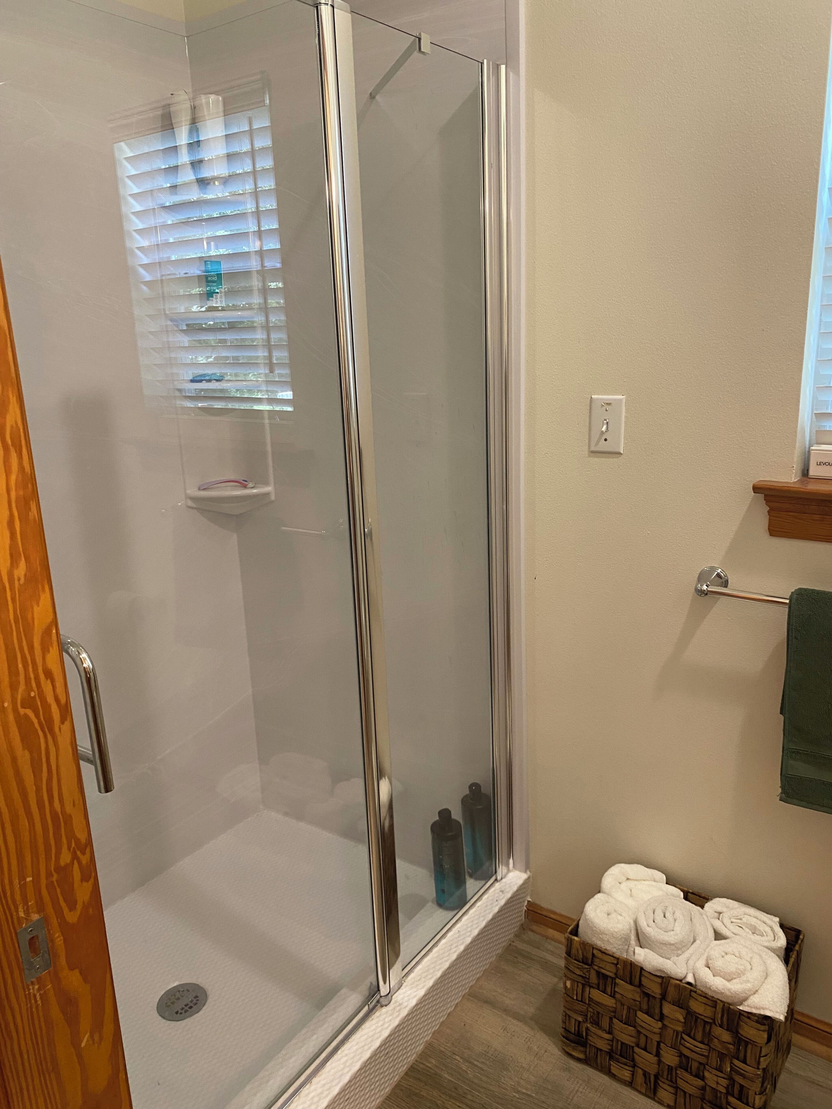 A glass-enclosed shower with a wooden door, a towel rack with a green towel, and a basket of white towels.