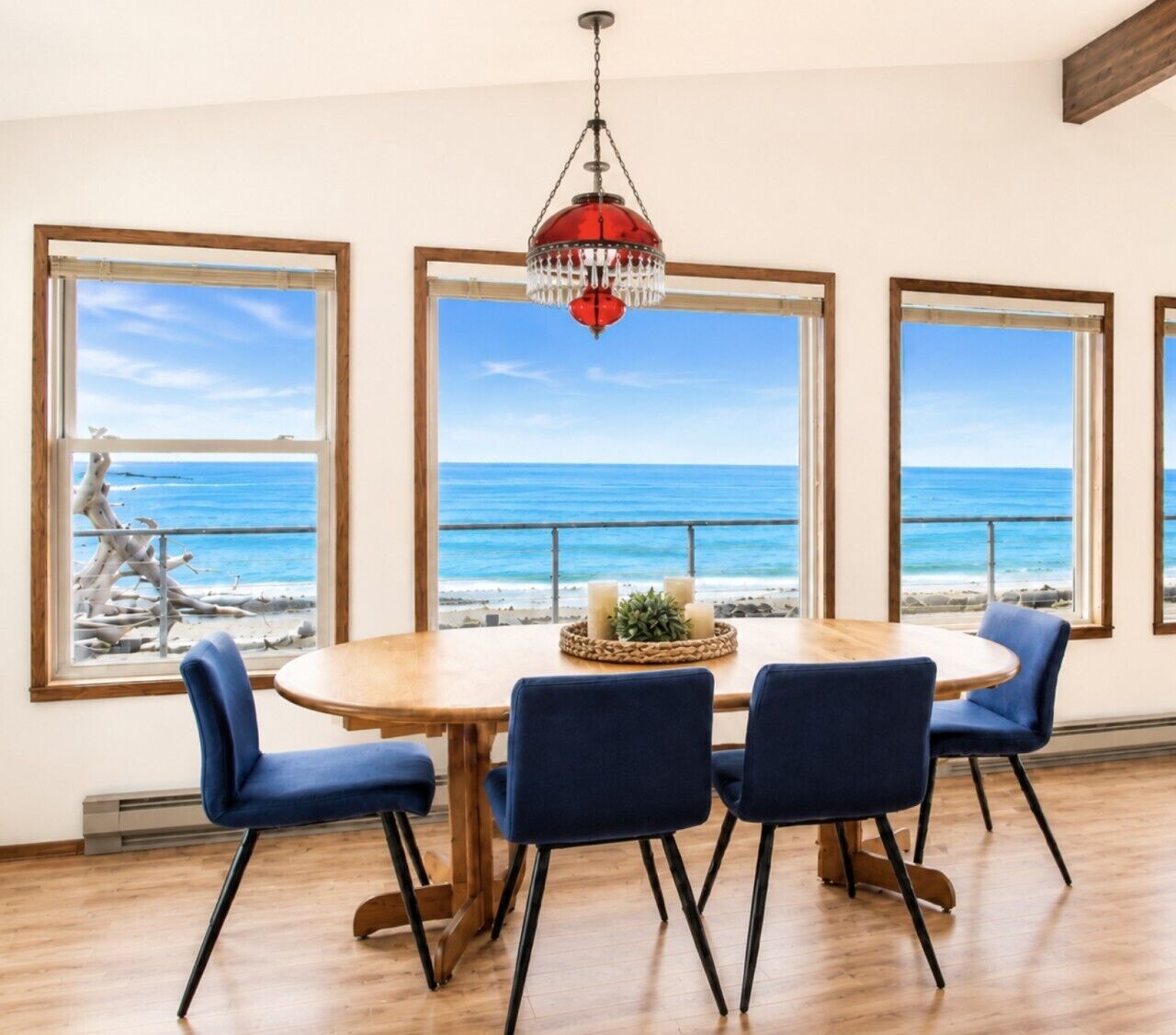 Un coin repas avec une table en bois et des chaises bleues, de grandes fenêtres donnant sur l’océan et un lustre rouge.