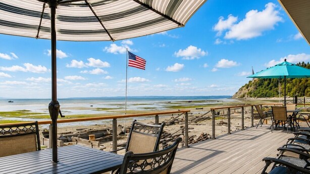 Terraza de madera con mobiliario exterior, con vista a una playa y una bandera.