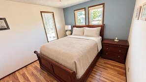 A bedroom with a wooden bed, a nightstand, a lamp, and a window with blinds.