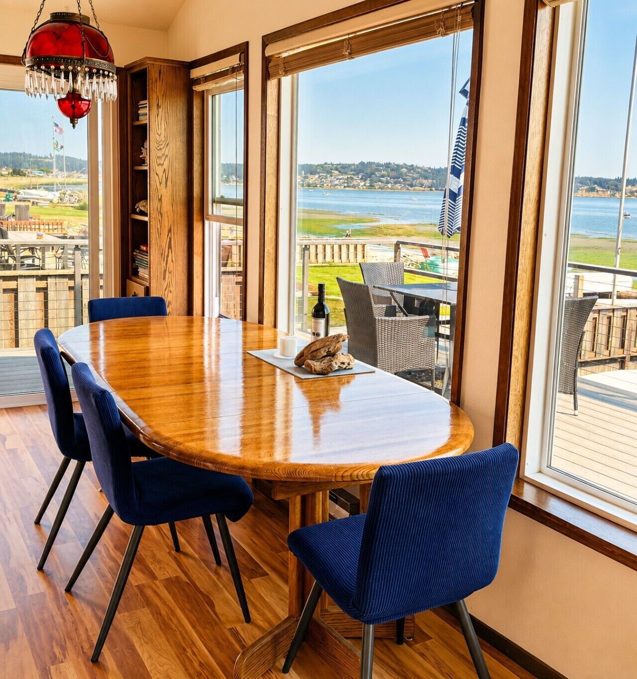 Un coin repas avec une table en bois et des chaises bleues, de grandes fenêtres donnant sur un plan d’eau et un quai.