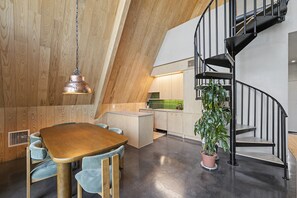 A modern kitchen with a wooden ceiling, a dining area with a wooden table and blue chairs, and a black spiral staircase.