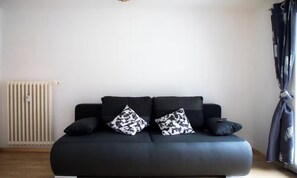 Um sofá preto com almofadas estampadas, um radiador e uma cortina.