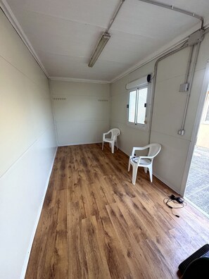 Ein Zimmer mit Holzboden, weißen Wänden, einem Fenster mit Metallgitter und zwei weißen Plastikstühlen.