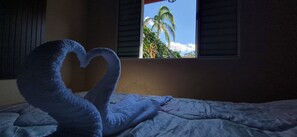 A bed with a heart-shaped pillow and a window with a view of palm trees. - Chalet at Sítio Bela Vista - 5 Minutes from the City Center (São Bento do Sapucaí)