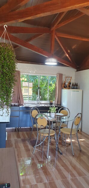 A kitchen with a dining area, wooden ceiling beams, a hanging plant, a window with curtains, and a refrigerator with bottles. - Chalet at Sítio Bela Vista - 5 Minutes from the City Center (São Bento do Sapucaí)