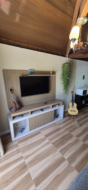 A living room with a wooden entertainment unit, a flat-screen TV, and a hanging plant. - Chalet at Sítio Bela Vista - 5 Minutes from the City Center (São Bento do Sapucaí)