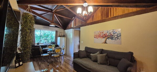 A cozy living area with a wooden ceiling, a sofa, a dining table with chairs, and a window with a view of greenery. - Chalet at Sítio Bela Vista - 5 Minutes from the City Center (São Bento do Sapucaí)