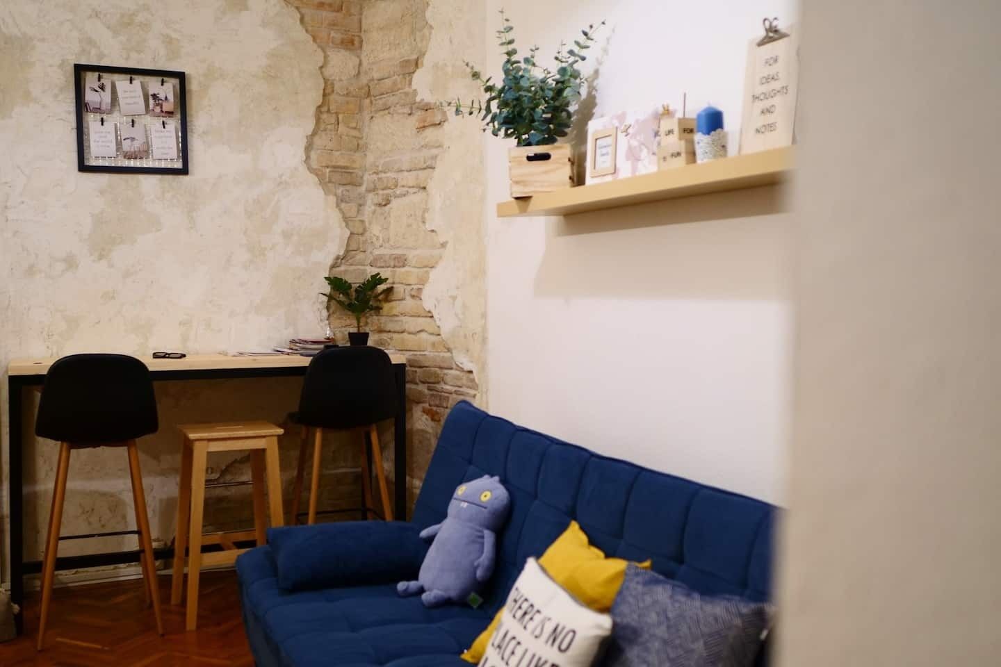 A room with a blue sofa, a small table, two chairs, a wall with a brick pattern, and a shelf with various items.