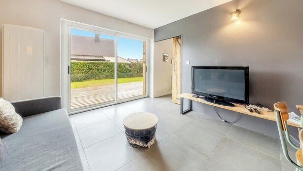 Una sala de estar moderna con un televisor de pantalla plana, un sofá, un otoman redóondo y un puerta corrediza de vidrio que da a un área exterior.