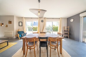 A dining area with a wooden table and chairs, a hanging light fixture, and a view through large windows. - Havre de Paix Familial à Deux Pas des Plages (Erdeven)