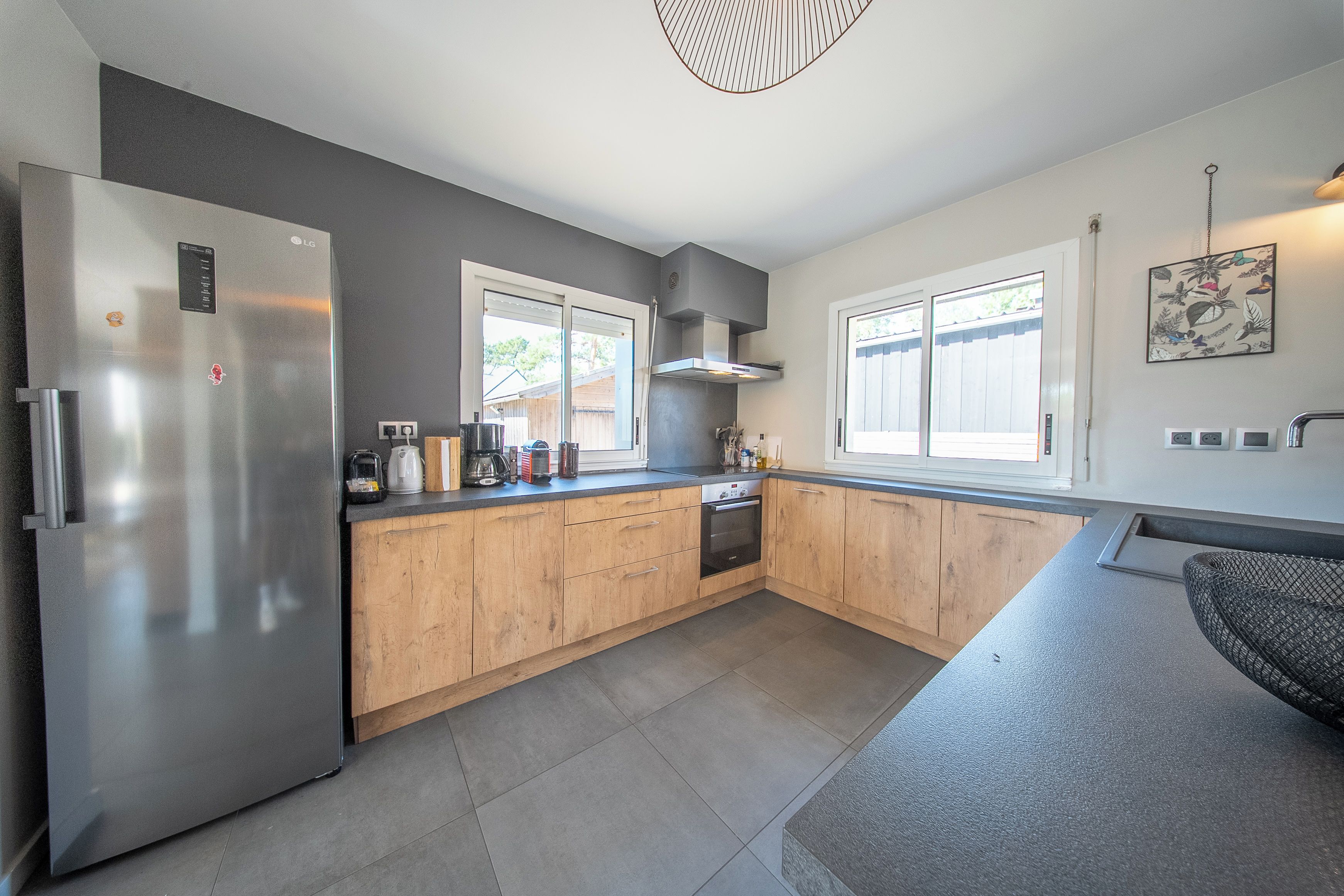 A modern kitchen with a stainless steel refrigerator, wooden cabinets, a built-in oven, and a window providing natural light.