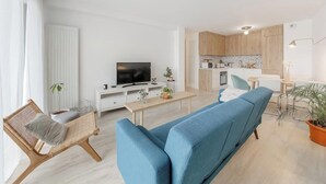 Una sala moderna con un sofá azul, una silla de mimbre, un televisor de pantalla plana y una cocina con armarios de madera.