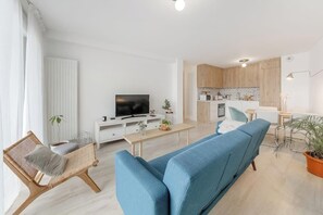 Ein modernes Wohnzimmer mit einem blauen Sofa, einem Korbsessel, einem Flachbildfernseher und einer Küchenzeile mit Holzausstattung.