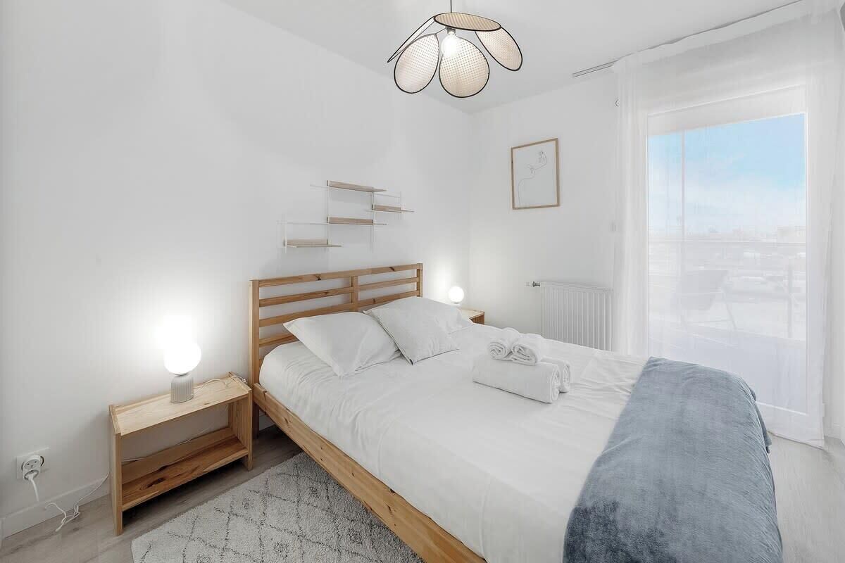 A modern bedroom with a wooden bed, bedside tables, a hanging light fixture, and a view of the city through a large window.