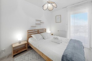 A modern bedroom with a wooden bed, bedside tables, a hanging light fixture, and a view of the city through a large window. - Appartement Neuf - Grande Terrasse & Parking Privé (Vannes)