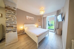 A bedroom with a bed, bedside lamps, a stone wall, a painting, a television, and a view of the outdoors. - La Rabine – Bel Appartement avec Terrasse Privée (Vannes)