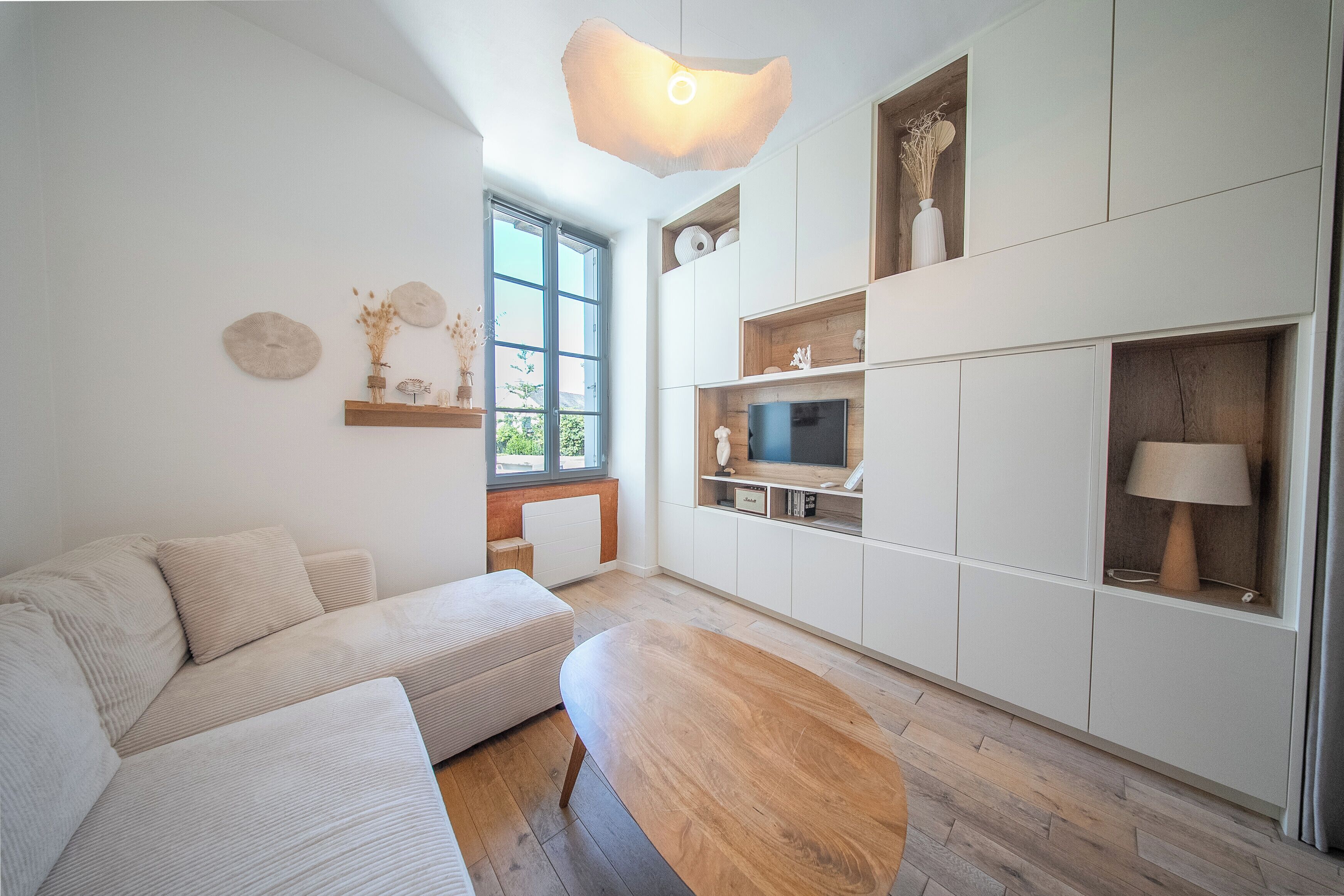 Una sala moderna con un sofá, una mesa de centro de madera, un televisor sobre una repisa de madera y una ventana con vistas a la vegetación.