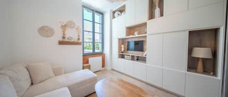 Una sala moderna con un sofá, una mesa de centro de madera, un televisor sobre una repisa de madera y una ventana con vistas a la vegetación.