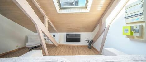 A room with a sloped ceiling, a skylight, a flat-screen TV, and a bed with white bedding.