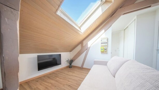 Una habitación con techo inclinado, un televisor de pantalla plana en la pared, un sofá blanco y suelo de madera.