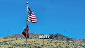 A flagpole with an American flag and a red flag with white text, set against a backdrop of a hill and a white building.