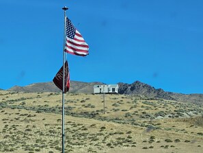 A flagpole with an American flag and a red flag with white text, set against a backdrop of a hill and a white building.