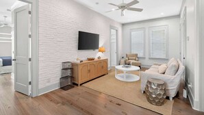 A modern living room with a white brick wall, a ceiling fan, a flat-screen TV, a wooden cabinet, a beige sofa, and a round coffee table. - Luxe Beach House: Steps to Rosemary, Beach, Pool & Bikes (Inlet Beach)