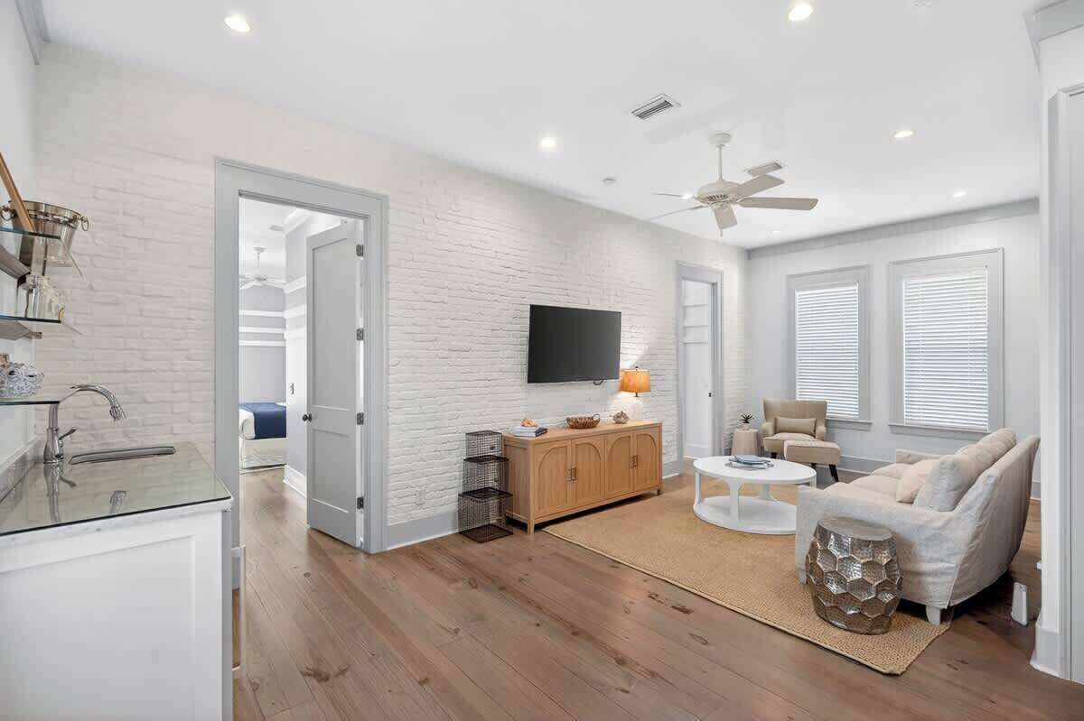 A modern living room with a white brick wall, a flat-screen TV, a ceiling fan, and a light-colored sofa.